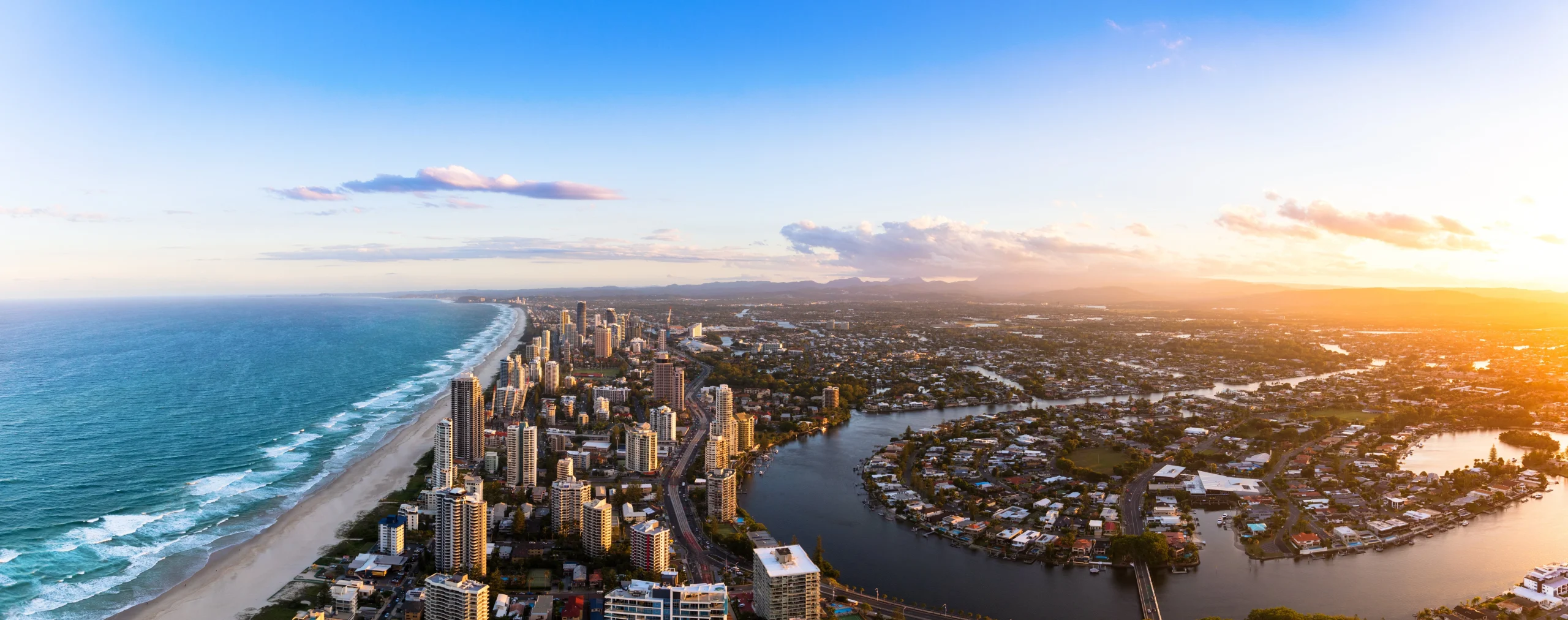 Aerial sunset panorama of Gold Coast Surfers Paradise skyscrapers lining a long sandy beach, with canal waterways and suburban sprawl stretching inland